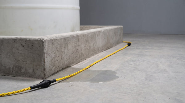 Câble jaune de détection de fuites posé le long d'une digue de rétention en béton, avec une flaque près d'un réservoir blanc